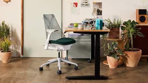 Ein Heimbüro mit einem Bambus-Schreibtisch und einem weißen Sayl-Stuhl mit grüner Sitzunterlage, umgeben von Terrakotta-Pflanzentöpfen