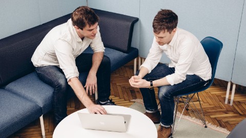 Two men look at a laptop in a Living Office Cove Setting. One is sitting on a blue Public Office Landscape couch and the other is sitting on a blue Eames Shell Chair.