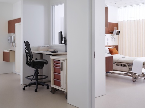 A caregiver workspace overlooking a patient room and equipped with a black Mirra 2 Stool, a height-adjustable table and mobile storage.