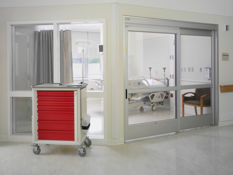 A mobile healthcare storage cart with casters and red drawers positioned outside a glass-walled patient room.