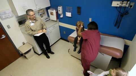 A nurse examines a child's ear while a man with a clipboard looks on.