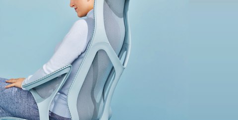 A woman wearing blue linen pants and a light blue sweater reclines in a Glacier light blue Cosm high-back ergonomic office chair.